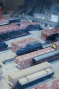 Model trains and tracks are displayed in a diorama, with some trains wrapped in patterned packaging. The scene includes a station building and various railway cars situated on tracks, creating a miniature railway setup.