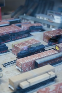 Model trains and tracks are displayed in a diorama, with some trains wrapped in patterned packaging. The scene includes a station building and various railway cars situated on tracks, creating a miniature railway setup.