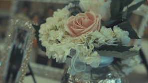 A close-up of a floral arrangement featuring a pink rose surrounded by white flowers and green leaves. The flowers are in a reflective, decorative vase, with blurred photo frames in the background adding a soft, elegant backdrop.