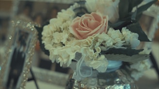 A close-up of a floral arrangement featuring a pink rose surrounded by white flowers and green leaves. The flowers are in a reflective, decorative vase, with blurred photo frames in the background adding a soft, elegant backdrop.