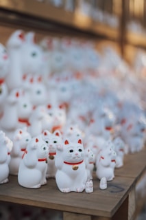 A collection of white ceramic cat figurines, known as maneki-neko, is neatly arranged on wooden display shelves. Each cat has a raised paw, red collars, and gold medallions around their necks. The background is softly blurred, giving focus to the rows of figurines in various sizes.