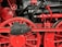 Close-up view of a red and black mechanical structure with large wheels, gears, and pipes. The intricate assembly showcases industrial engineering with detailed components and metallic textures.