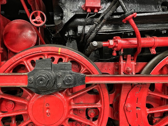 Close-up view of a red and black mechanical structure with large wheels, gears, and pipes. The intricate assembly showcases industrial engineering with detailed components and metallic textures.