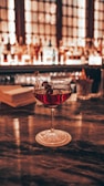 A cocktail glass filled with a red drink garnished with dark berries on a skewer sits on a coaster on a polished bar counter, with a blurred background of a backlit bar shelf stocked with various bottles.