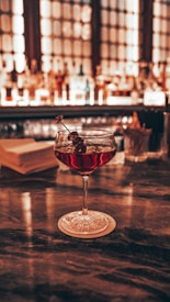 A cocktail glass filled with a red drink garnished with dark berries on a skewer sits on a coaster on a polished bar counter, with a blurred background of a backlit bar shelf stocked with various bottles.