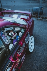 A customized car with vibrant decals and stickers on the side, including various anime characters and text. The vehicle features a shiny magenta paint, white wheels, and a rear wing, parked on a gray asphalt surface next to a chain-link fence.