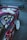 A customized car with vibrant decals and stickers on the side, including various anime characters and text. The vehicle features a shiny magenta paint, white wheels, and a rear wing, parked on a gray asphalt surface next to a chain-link fence.