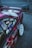 A customized car with vibrant decals and stickers on the side, including various anime characters and text. The vehicle features a shiny magenta paint, white wheels, and a rear wing, parked on a gray asphalt surface next to a chain-link fence.