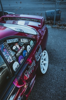 A customized car with vibrant decals and stickers on the side, including various anime characters and text. The vehicle features a shiny magenta paint, white wheels, and a rear wing, parked on a gray asphalt surface next to a chain-link fence.