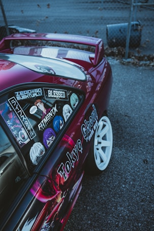 A customized car with vibrant decals and stickers on the side, including various anime characters and text. The vehicle features a shiny magenta paint, white wheels, and a rear wing, parked on a gray asphalt surface next to a chain-link fence.