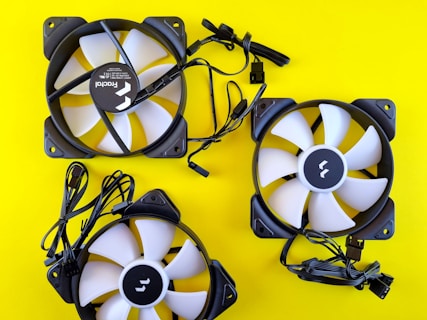 Three computer cooling fans with black frames and white blades are arranged on a bright yellow background. Each fan has a label at the center and is connected to a black cable ending with a connector.