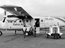 A small aircraft with the text 'Tara Air' on its side is parked on an airport runway. The plane has a high wing and twin turboprop engines. Three individuals in work attire are gathered near the plane's entrance, while a ground power unit is connected to the aircraft.