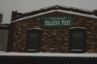 A brick building facade with a sign reading 'Cloudcroft Trading Post' and 'Beads-Rocks-Jewelry' under it. The structure has a snow-covered roof, and two windows bordered by dark trim on either side of the sign.