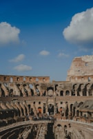 The image features the ancient Colosseum in Rome, with its iconic arches and elliptical structure. A clear blue sky with scattered clouds forms the backdrop. The amphitheater's weathered stone and brickwork are highlighted, with tourists gathered around its interior, providing a sense of scale to the historical monument.