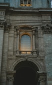 An architectural facade featuring a large arched window with intricate details and stone columns. The window is illuminated by a warm light, creating a contrast against the grey stone structure. Ornamental stone carvings are present above and below the window, with the overall style reflecting classical or Renaissance influences.