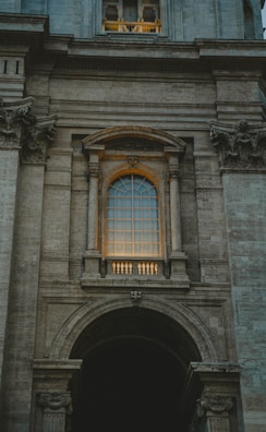 An architectural facade featuring a large arched window with intricate details and stone columns. The window is illuminated by a warm light, creating a contrast against the grey stone structure. Ornamental stone carvings are present above and below the window, with the overall style reflecting classical or Renaissance influences.