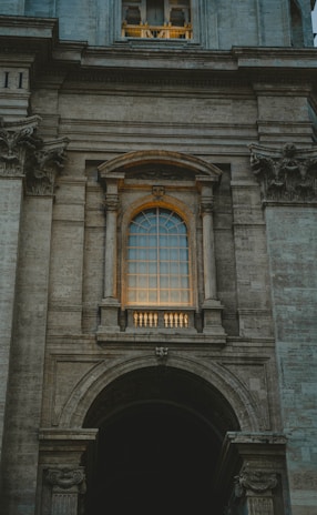 An architectural facade featuring a large arched window with intricate details and stone columns. The window is illuminated by a warm light, creating a contrast against the grey stone structure. Ornamental stone carvings are present above and below the window, with the overall style reflecting classical or Renaissance influences.