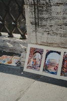 Three watercolor paintings are propped against a textured concrete wall. The artwork features vibrant street scenes, including an archway and the Colosseum, suggesting an Italian cityscape. A palette with paint, brushes, and a jar with water sits nearby on the ground, indicating an outdoor painting session.