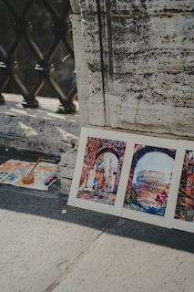 Three watercolor paintings are propped against a textured concrete wall. The artwork features vibrant street scenes, including an archway and the Colosseum, suggesting an Italian cityscape. A palette with paint, brushes, and a jar with water sits nearby on the ground, indicating an outdoor painting session.