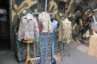 Several mannequins display military-style jackets and shirts with various camouflage patterns. The background features a wall painted in a mix of camouflage colors. The scene includes elements like a hand truck and a broom, suggesting a setting outside a military surplus store.