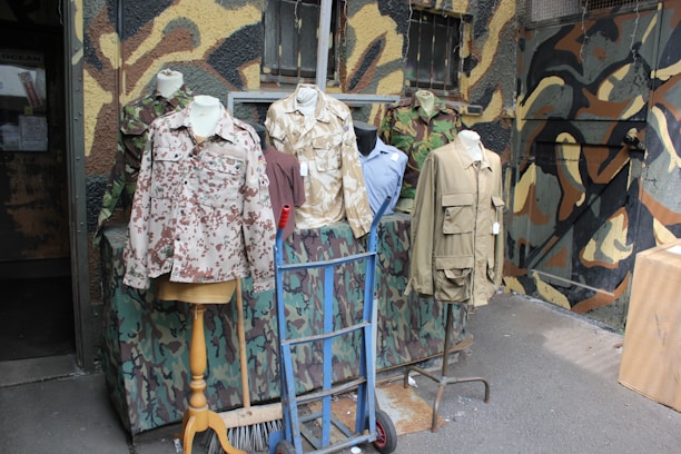 Several mannequins display military-style jackets and shirts with various camouflage patterns. The background features a wall painted in a mix of camouflage colors. The scene includes elements like a hand truck and a broom, suggesting a setting outside a military surplus store.