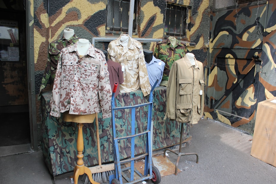 Several mannequins display military-style jackets and shirts with various camouflage patterns. The background features a wall painted in a mix of camouflage colors. The scene includes elements like a hand truck and a broom, suggesting a setting outside a military surplus store.