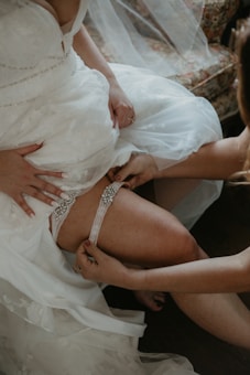 A bride is adjusting her white lace dress while another person helps her with a decorative garter. The setting includes elegant white fabric with floral patterns.