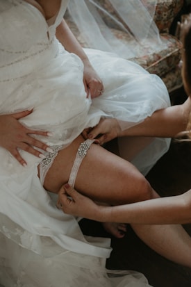 A bride is adjusting her white lace dress while another person helps her with a decorative garter. The setting includes elegant white fabric with floral patterns.