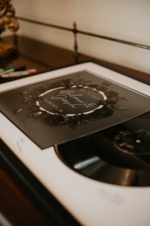 A framed circular print with floral decorations is resting on a piece of furniture next to some colored pens. The print includes cursive text and details like dates and locations, giving it an elegant and personalized appearance. The lighting is warm, creating a cozy atmosphere.