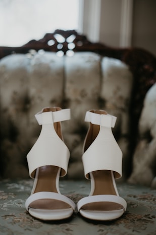 Two elegant white high-heeled sandals with thick ankle straps are placed on a floral upholstered vintage sofa. The background is softly blurred, highlighting the shoes.