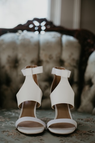 Two elegant white high-heeled sandals with thick ankle straps are placed on a floral upholstered vintage sofa. The background is softly blurred, highlighting the shoes.