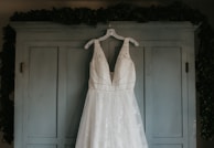 A white wedding dress with intricate lace detailing hangs from a white hanger on the doors of a light blue armoire. The background is subtly adorned with leafy greenery draped over the top edges of the armoire.