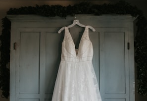 A white wedding dress with intricate lace detailing hangs from a white hanger on the doors of a light blue armoire. The background is subtly adorned with leafy greenery draped over the top edges of the armoire.