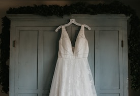A white wedding dress with intricate lace detailing hangs from a white hanger on the doors of a light blue armoire. The background is subtly adorned with leafy greenery draped over the top edges of the armoire.