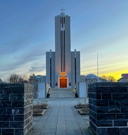 A towering church stands prominently at the end of a paved pathway, framed by dark stone walls. The building is minimalist and modern, with vertical black accents running up its facade topped by a cross. The entrance is warmly lit, creating a contrast with the cool blue and gray tones of the twilight sky. Sparse vegetation lines the walkway, leading the eye toward the structure.