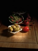 A wooden surface holds a small white bowl filled with seasoned green olives. Nearby, ripe red tomatoes rest on the surface. In the background, there is a bowl containing a salad of leafy greens, sprouts, and more tomatoes, partially obscured by shadows.
