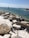 A rocky shoreline with large stones scattered on white sandy ground. The ocean stretches out in the background, with blue and green hues. Two channel markers, one with a red triangle and one with a green square top, are positioned in the water, guiding nearby boats.