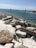 A rocky shoreline with large stones scattered on white sandy ground. The ocean stretches out in the background, with blue and green hues. Two channel markers, one with a red triangle and one with a green square top, are positioned in the water, guiding nearby boats.