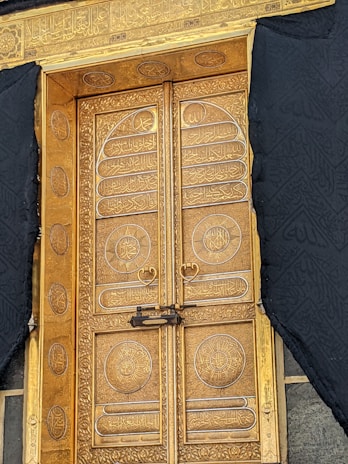 A beautifully ornate golden door adorned with intricate Arabic calligraphy and decorative patterns. The door is framed by black drapery featuring subtle designs, enhancing its elegance. The door has heart-shaped handles and is secured by a visible lock.
