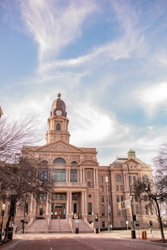 A grand, historic courthouse building with a clock tower, featuring neoclassical architectural elements, stands prominently against a bright sky with wispy clouds. Tall trees with bare branches frame the scene, indicating a late autumn or winter setting.