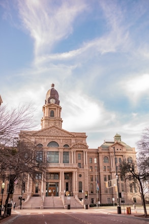 A grand, historic courthouse building with a clock tower, featuring neoclassical architectural elements, stands prominently against a bright sky with wispy clouds. Tall trees with bare branches frame the scene, indicating a late autumn or winter setting.
