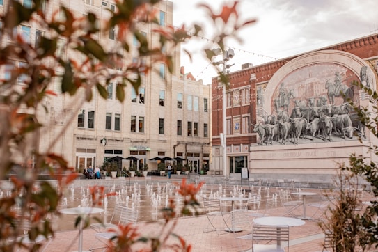 A public square featuring outdoor seating and a decorative water fountain, surrounded by historic brick buildings. The background displays a large mural depicting cattle, with a few people visible near a caf&eacute;.