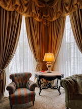 A luxurious interior corner featuring heavy golden drapes and lace curtains allowing soft light to filter through. An ornate table with a lamp and an open book rests between an elegant, richly-upholstered armchair and a plush green sofa. The intricate patterns on the carpet complement the opulent decor.