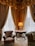 A luxurious interior corner featuring heavy golden drapes and lace curtains allowing soft light to filter through. An ornate table with a lamp and an open book rests between an elegant, richly-upholstered armchair and a plush green sofa. The intricate patterns on the carpet complement the opulent decor.