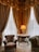 A luxurious interior corner featuring heavy golden drapes and lace curtains allowing soft light to filter through. An ornate table with a lamp and an open book rests between an elegant, richly-upholstered armchair and a plush green sofa. The intricate patterns on the carpet complement the opulent decor.