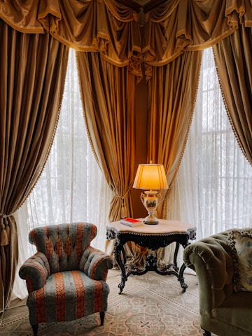 A luxurious interior corner featuring heavy golden drapes and lace curtains allowing soft light to filter through. An ornate table with a lamp and an open book rests between an elegant, richly-upholstered armchair and a plush green sofa. The intricate patterns on the carpet complement the opulent decor.