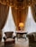 A luxurious interior corner featuring heavy golden drapes and lace curtains allowing soft light to filter through. An ornate table with a lamp and an open book rests between an elegant, richly-upholstered armchair and a plush green sofa. The intricate patterns on the carpet complement the opulent decor.