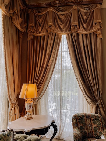 An ornate interior setting with luxurious draped curtains on a large window, a table with an elegant lamp featuring a decorative base, and a plush upholstered chair. The curtains are richly textured, and the lamp is illuminated, casting a warm glow.