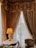 An ornate interior setting with luxurious draped curtains on a large window, a table with an elegant lamp featuring a decorative base, and a plush upholstered chair. The curtains are richly textured, and the lamp is illuminated, casting a warm glow.