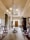 An elegant, vintage hallway features ornate wooden furniture, including armchairs with pink upholstery and a central round table. The room is adorned with a grand chandelier hanging from the ceiling. The walls are decorated with framed pictures and a large mirror. There is rich woodwork, with cabinets and a tall grandfather clock situated near the closed double doors at the end of the hallway. A long decorative rug runs along the wooden floor.